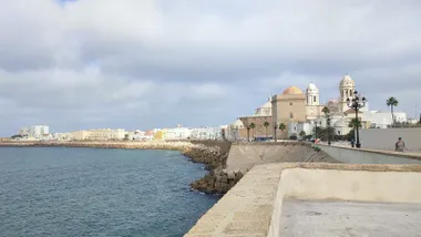 Imagen del Campo del Sur en Cádiz con cielo cubierto de nubes grises. Casi en primer plano, la Catedral, en contraste con el mar en calma.