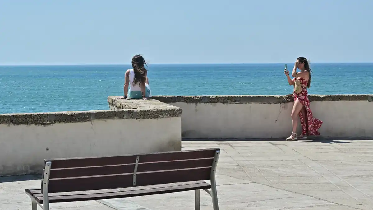 En la balaustrada de piedra del Campo del Sur, una turista viste ropa ligera de verano y posa mirando hacia la cámara, mientras su acompañante, de espaldas, le toma una foto. El viento de Levante mueve sus ropas y cabellos; detrás, el mar azul intenso y el cielo claro completan la escena.