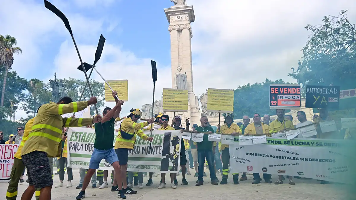 Momento de la manifestación de los bomberos forestales del Plan Infoca en Cádiz. Varios trabajadores con ropa de intervención sostienen pancartas con lemas como “Se vende Infoca” y “Nos ponen en peligro”, mientras el humo rojo y verde cubre parte de la Plaza de España, frente al monumento de la Constitución de 1812.