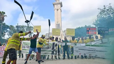 Momento de la manifestación de los bomberos forestales del Plan Infoca en Cádiz. Varios trabajadores con ropa de intervención sostienen pancartas con lemas como “Se vende Infoca” y “Nos ponen en peligro”, mientras el humo rojo y verde cubre parte de la Plaza de España, frente al monumento de la Constitución de 1812.