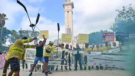 Momento de la manifestación de los bomberos forestales del Plan Infoca en Cádiz. Varios trabajadores con ropa de intervención sostienen pancartas con lemas como “Se vende Infoca” y “Nos ponen en peligro”, mientras el humo rojo y verde cubre parte de la Plaza de España, frente al monumento de la Constitución de 1812.