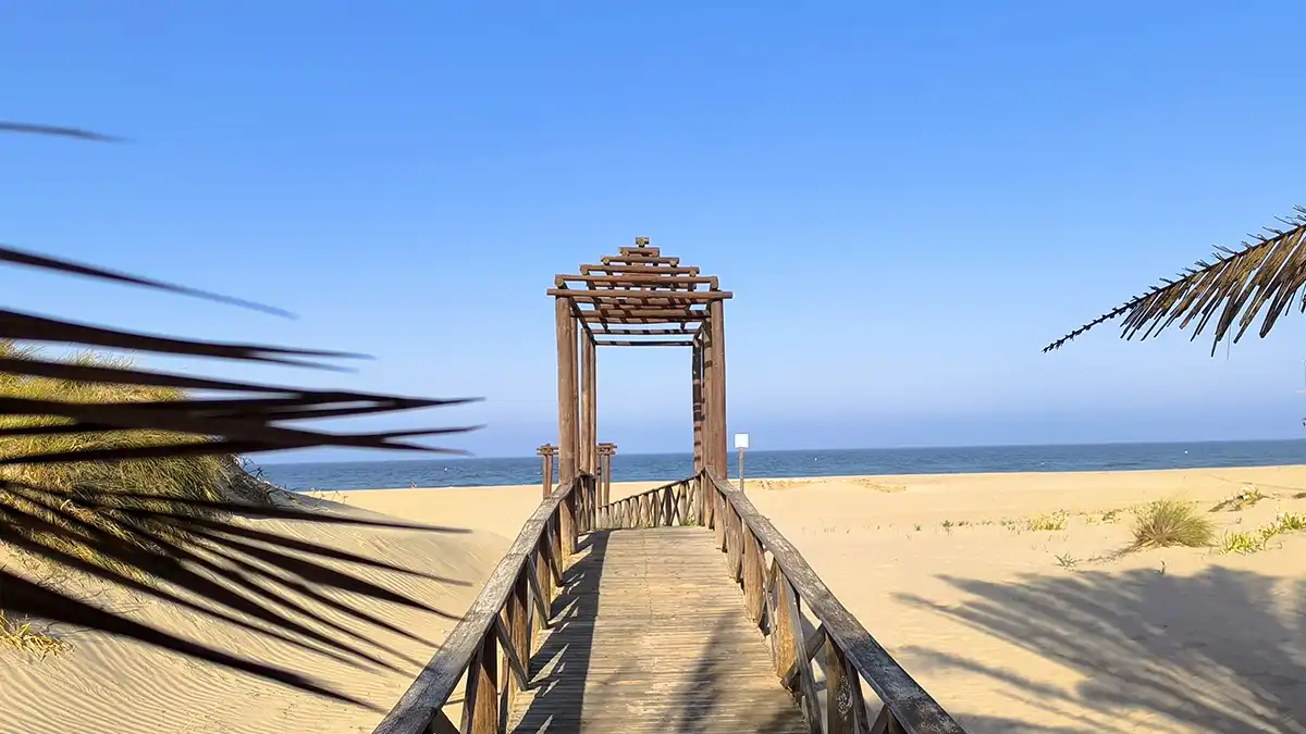Vista de una pasarela de madera que conduce hasta la arena de la playa de Cortadura, con el mar al fondo.