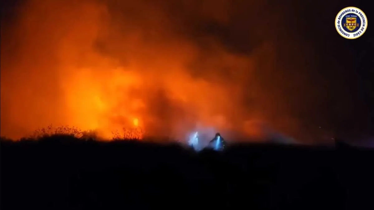 Llamas destacando sobre la oscuridad y un bombero con linterna y luz azulada.