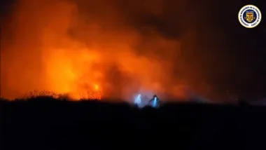 Llamas destacando sobre la oscuridad y un bombero con linterna y luz azulada.