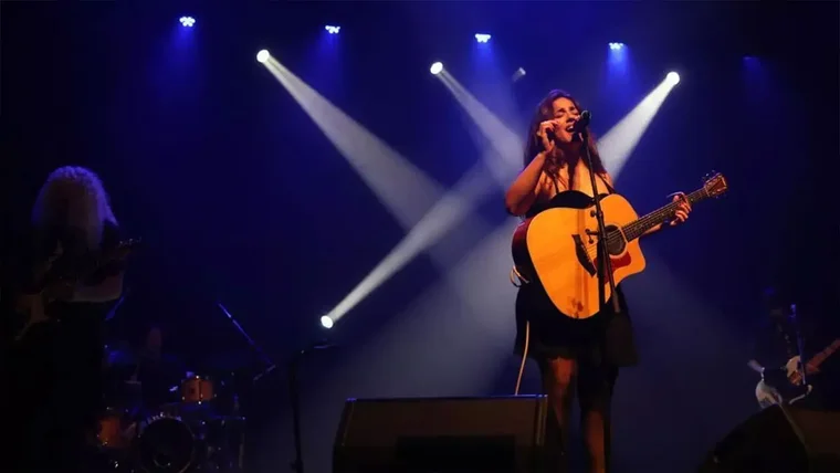 Julia Medina sosteniendo una guitarra en el escenario y con focos blancos y azules tras ella.