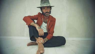 El músico Leiva sentado en el suelo, descalzo, con camisa roja y sombrero.
