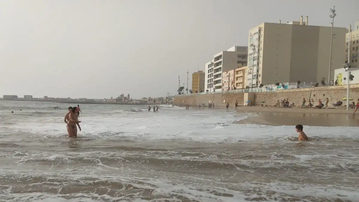 Imagen de un niño que ha sido arrastrado por una ola en la orilla de la Playa de la Victoria. A su lado, un adulto le advierte del peligro. De fondo se aprecia la Catedral de Cádiz y el mar agitado.