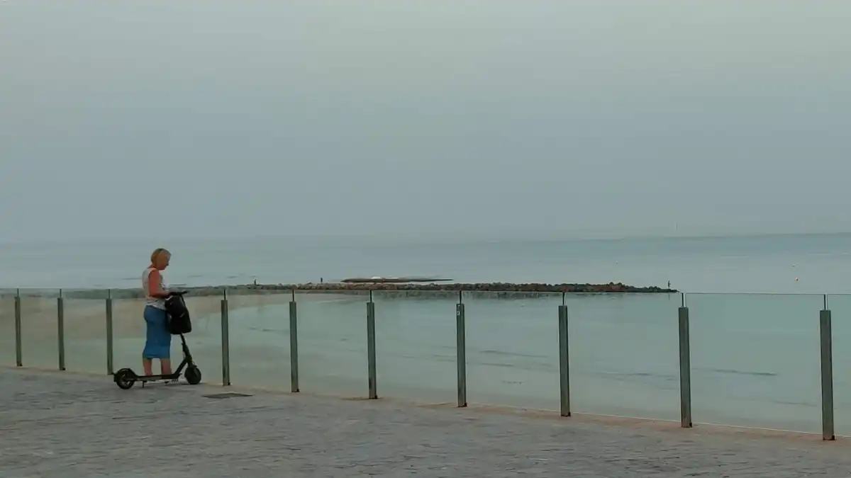 Una mujer en patinete se detiene junto a la barandilla de cristal del paseo de la playa de Santa María del Mar de Cádiz, mirando hacia el mar. Al fondo, el cielo está blanquecino por la bruma y la humedad, reflejando el ambiente sofocante típico de los días de bochorno.