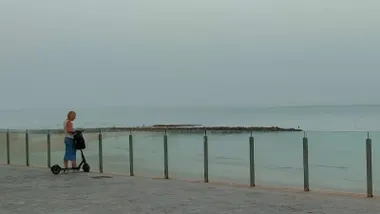 Una mujer en patinete se detiene junto a la barandilla de cristal del paseo de la playa de Santa María del Mar de Cádiz, mirando hacia el mar. Al fondo, el cielo está blanquecino por la bruma y la humedad, reflejando el ambiente sofocante típico de los días de bochorno.