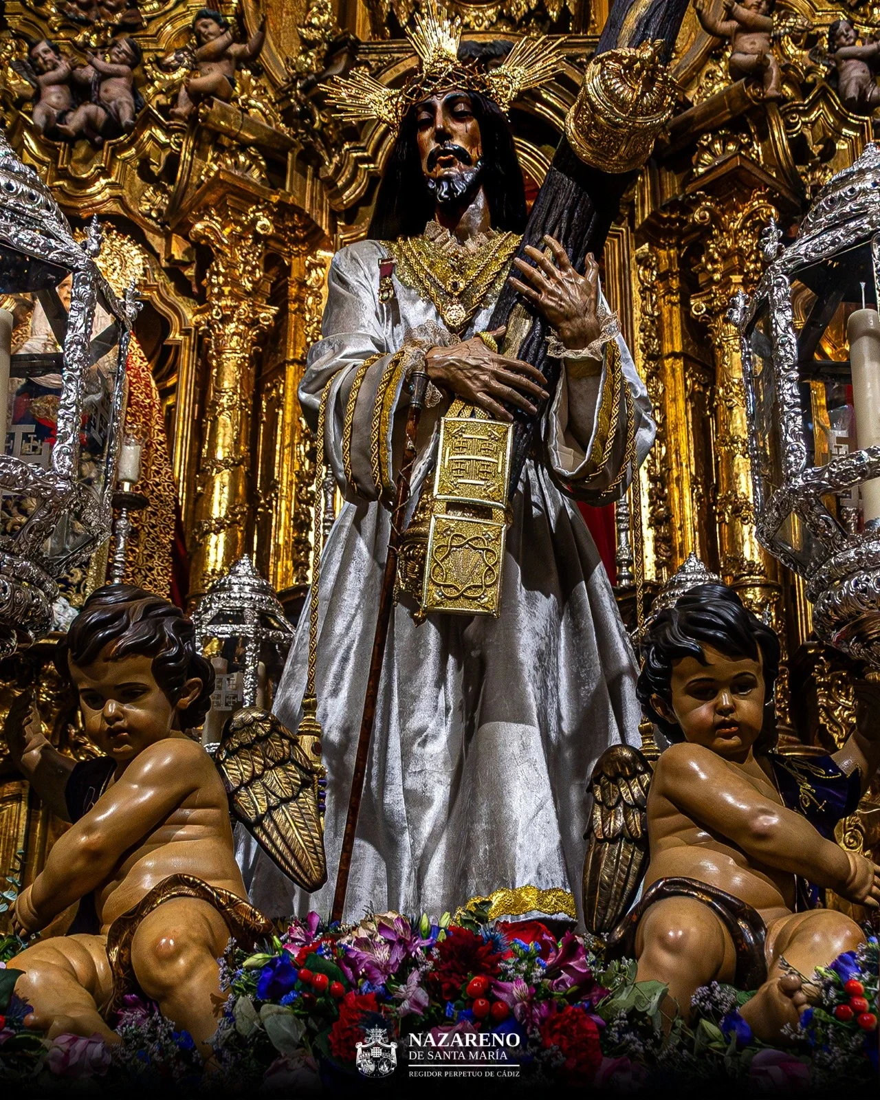 Imagen del Nazareno de Santa María sobre la parihuela en la que realizará la peregrinación y ya preparada con el exorno floral y los angelotes. El Regidor Perpetuo viste la túnica color perla que supone la gran novedad de esta peregrinación.