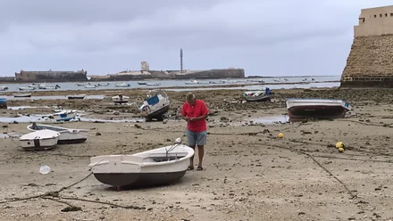 Vista de la playa de La Caleta en bajamar, con barcas sobre las rocas y un hombre trabajando en una de ellas. Al fondo, el castillo de San Sebastián y, a la derecha, el de Santa Catalina. El cielo aparece nublado, con una luz húmeda que acentúa el ambiente de bochorno