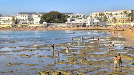Imagen de la playa de La Caleta en Cádiz durante la tarde, con la marea muy baja que deja al descubierto gran parte de la arena húmeda. Muchas personas disfrutan de la jornada tanto en la orilla como en el agua. Al fondo se aprecia el histórico Balneario de La Palma, con el cielo despejado que refuerza el ambiente veraniego de la escena.