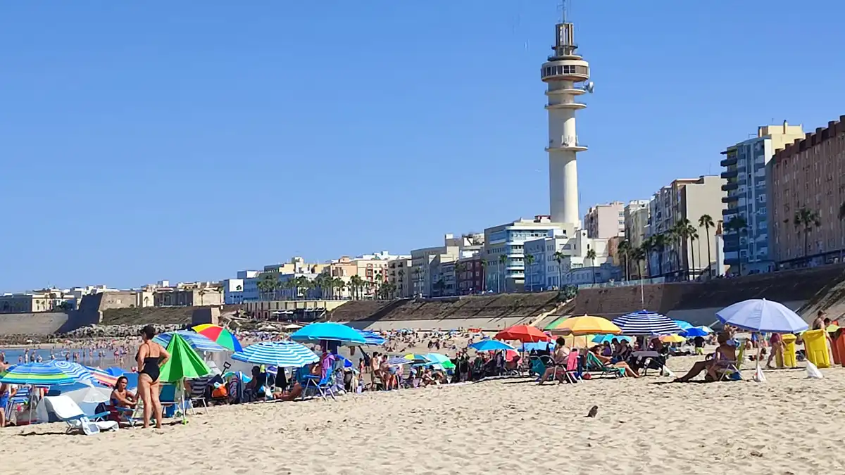 Vista de la playa de Santa María del Mar, en Cádiz, con numerosos bañistas en la arena y en el agua. El cielo está despejado, en un día caluroso con viento de Levante