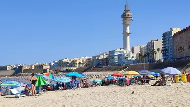 Vista de la playa de Santa María del Mar, en Cádiz, con numerosos bañistas en la arena y en el agua. El cielo está despejado, en un día caluroso con viento de Levante