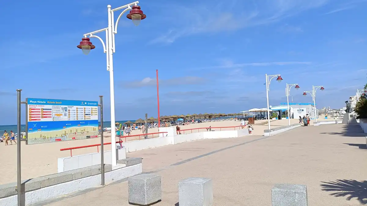 Fotografía tomada desde el paseo marítimo de Cádiz, donde se observa la playa de la Victoria con el mar en calma, arena clara y un cielo completamente azul y despejado.