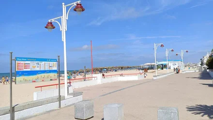 Fotografía tomada desde el paseo marítimo de Cádiz, donde se observa la playa de la Victoria con el mar en calma, arena clara y un cielo completamente azul y despejado.