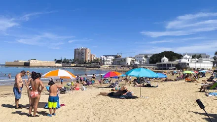 Imagen de la playa de la Caleta en Cádiz con bastante público. En primer plano, a la izquierda, una familia se aplica crema solar, y al fondo a la derecha se ve el Balneario de la Palma.