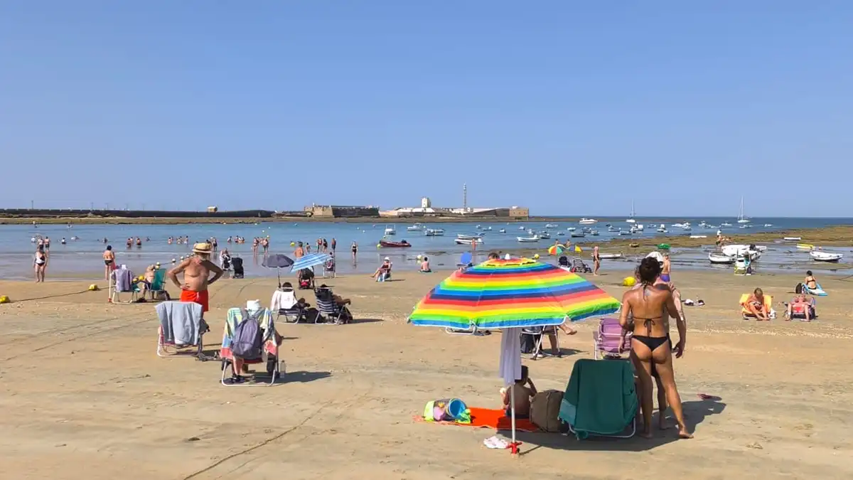 Mujer de espaldas en la arena de la playa de La Caleta, Cádiz, aplicando crema solar en la espalda de un hombre sentado frente al mar, con cielo azul despejado y bañistas al fondo.