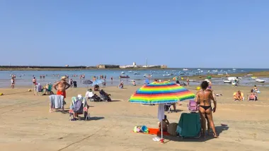 Mujer de espaldas en la arena de la playa de La Caleta, Cádiz, aplicando crema solar en la espalda de un hombre sentado frente al mar, con cielo azul despejado y bañistas al fondo.