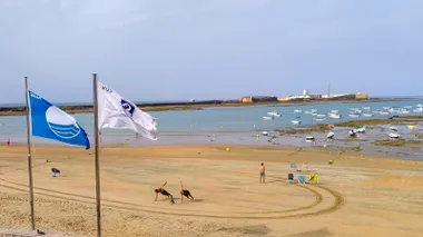 Imagen de la playa de la Caleta en Cádiz en la mañana del 26 de agosto. El cielo está algo nublado y con un poco de bruma, transmitiendo sensación de frescor. En primer plano, una pareja practica deporte sobre la arena húmeda en un ambiente tranquilo y sereno