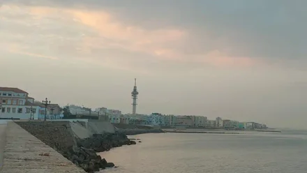Panorámica tomada desde el Campo del Sur en Cádiz: se ven las playas de Santa María del Mar y la Victoria bajo un cielo con nubes y notable humedad; al fondo destaca la torre de comunicaciones (“pirulí”) sobre el perfil urbano.