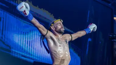 El luchador gaditano Carlos Coello, con pantalón de Muay Thai y guantes blancos, levanta los brazos en alto durante su presentación en el ring del Estadio Rajadamnern en Bangkok, ante un público expectante.