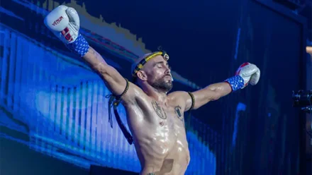 El luchador gaditano Carlos Coello, con pantalón de Muay Thai y guantes blancos, levanta los brazos en alto durante su presentación en el ring del Estadio Rajadamnern en Bangkok, ante un público expectante.