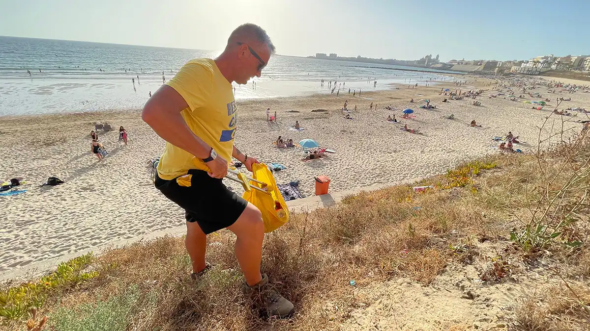Quique Bolsitas, de perfil, mientras recoge colillas y pequeños residuos en la playa de Santa María del Mar, en Cádiz. Lleva guantes y sostiene una bolsa de basura parcialmente llena.