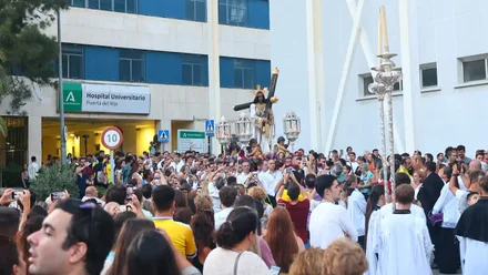 El Nazareno de Santa María, con túnica perla y cruz arbórea, sale del Hospital Puerta del Mar rodeado de fieles y acompañado por el cartel del centro hospitalario al fondo, mientras decenas de personas lo reciben en la calle.