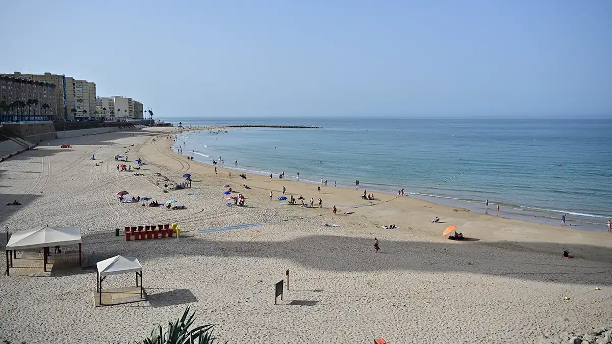Vista de la playa de Santa María del Mar en Cádiz, con numerosos bañistas desde primera hora de la mañana, buscando alivio del calor en plena ola de calor. El cielo aparece despejado y la bruma del bochorno es visible en el horizonte.