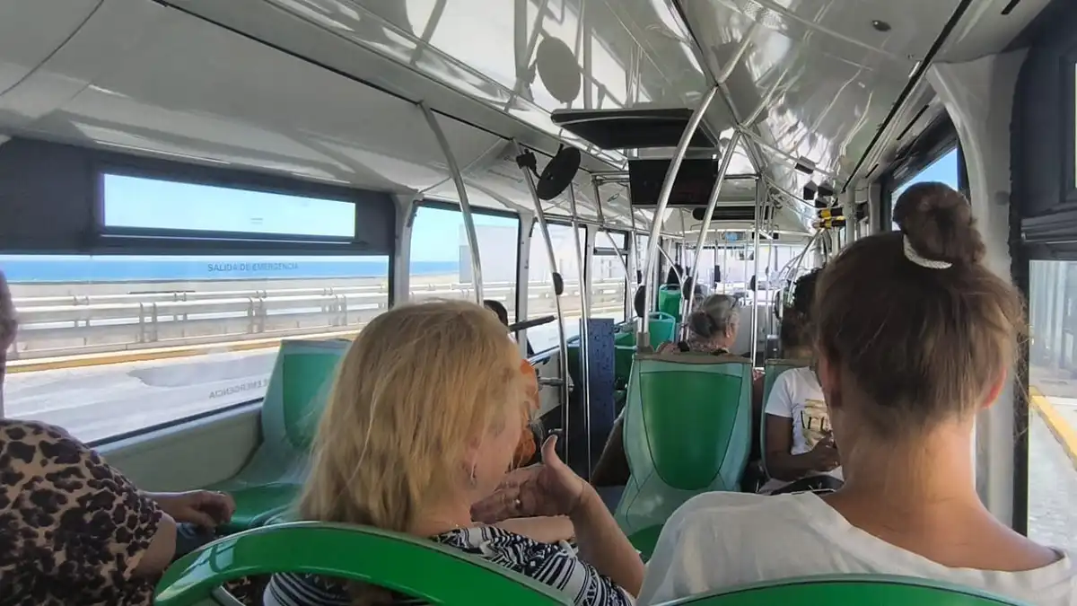 Pasajeros sentados dentro de un autobús urbano se abanican con la mano para aliviar el calor; la luz del sol entra por las ventanillas.