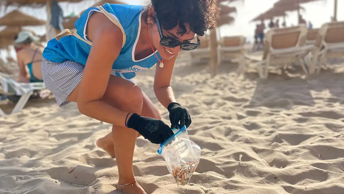 Una coluntaria, agachada, recoge colillas de la arena de la Playa de Santa María del Mar.