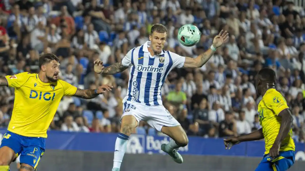 Dos jugadores, uno del Leganés con camiseta blanca y otro del Cádiz con camiseta oscura, saltan por un balón que aparece teñido de verde fluorescente.