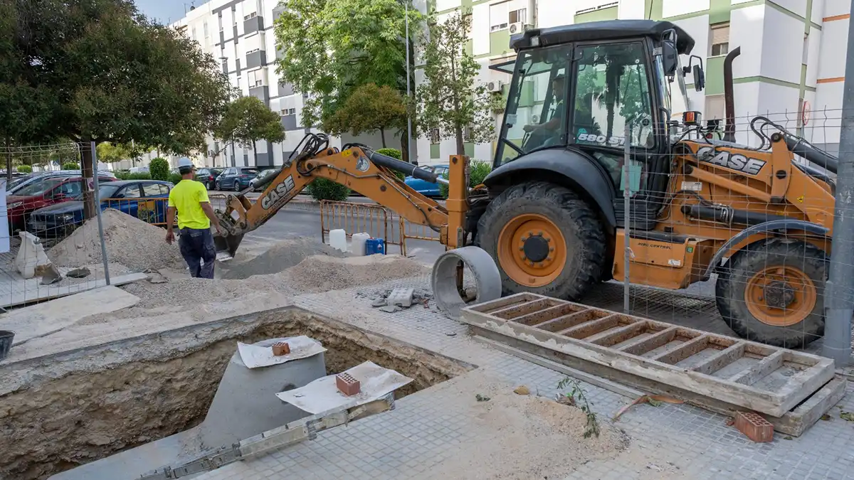 Varios operarios con chalecos reflectantes colocan nuevo pavimento en una acera de Cádiz, junto a maquinaria y señalización de obras.
