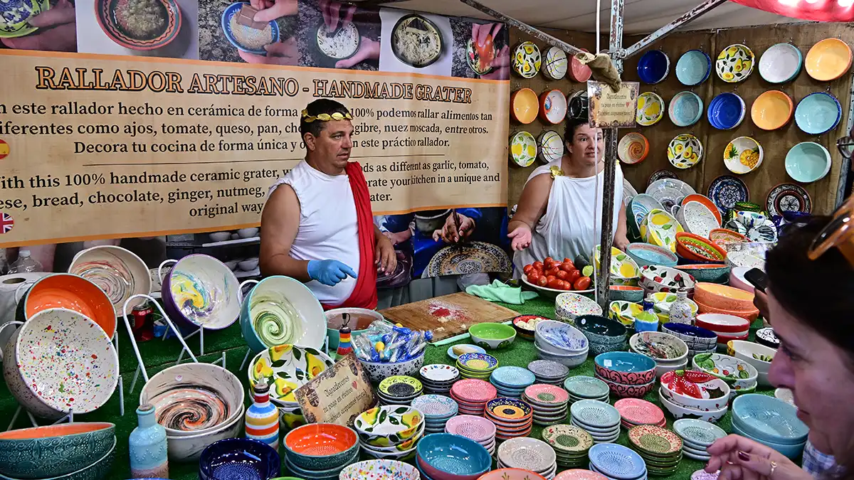 Hombre y mujer con túnicas rojas y capas púrpuras atienden en su puesto de alfarería de este Mercado Romano