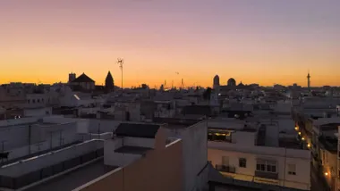 Vista de un amanecer en Cádiz: el cielo teñido de tonos anaranjados y rosados ilumina las siluetas de iglesias y casas en primer plano.