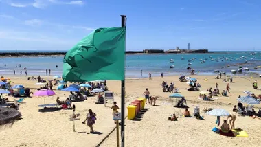 Vista de la playa de la Caleta en Cádiz con una bandera verde ondeando en el mástil por el viento de poniente. En segundo plano, el castillo de San Sebastián se recorta frente al mar con oleaje moderado.