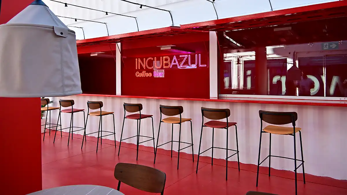 Espacio interior con mesas redondas, sillones y una barra de cafetería. Emprendedores conversan y comparten café en un ambiente informal.
