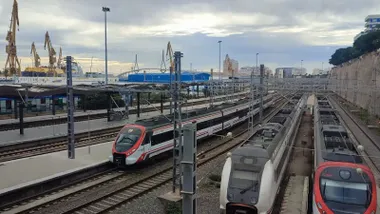 Vista de la estación de trenes de Cádiz con varios convoyes detenidos en las vías exteriores, bajo un cielo cubierto de nubes grises; al fondo se distinguen las grúas de los astilleros.