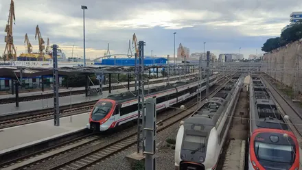 Vista de la estación de trenes de Cádiz con varios convoyes detenidos en las vías exteriores, bajo un cielo cubierto de nubes grises; al fondo se distinguen las grúas de los astilleros.