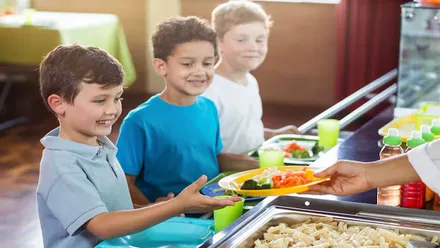 Tres niños en un comedor escolar, le dan platos con el menú.