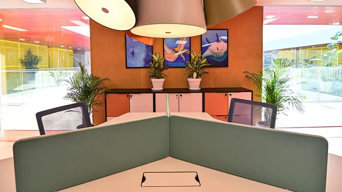 Sala luminosa con mesas compartidas, sillas ergonómicas y paredes acristaladas. Varias personas trabajan con portátiles en un entorno abierto y colaborativo.