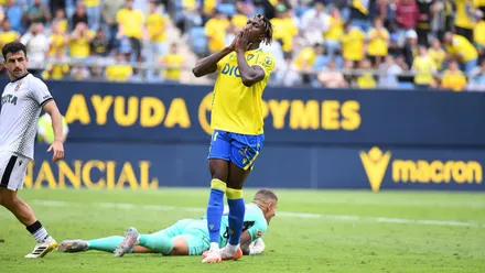 Dawda, futbolista del Cádiz CF, aparece en primer plano con gesto de frustración tras fallar una ocasión clara. Se echa las manos a la cara mientras el portero rival está en el suelo con el balon atrapado de fondo.