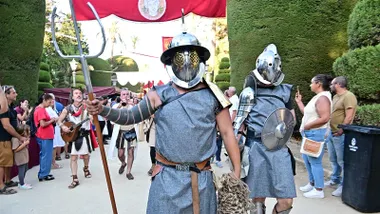 Dos actores vestidos de gladiadores romanos, con cascos metálicos, escudos y espadas, participan en el pasacalles del Mercado Romano que recorre el Parque Genovés. A su alrededor, visitantes siguen la comitiva y fotografían la recreación histórica.