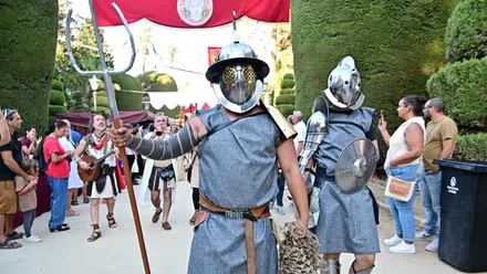 Dos actores vestidos de gladiadores romanos, con cascos metálicos, escudos y espadas, participan en el pasacalles del Mercado Romano que recorre el Parque Genovés. A su alrededor, visitantes siguen la comitiva y fotografían la recreación histórica.