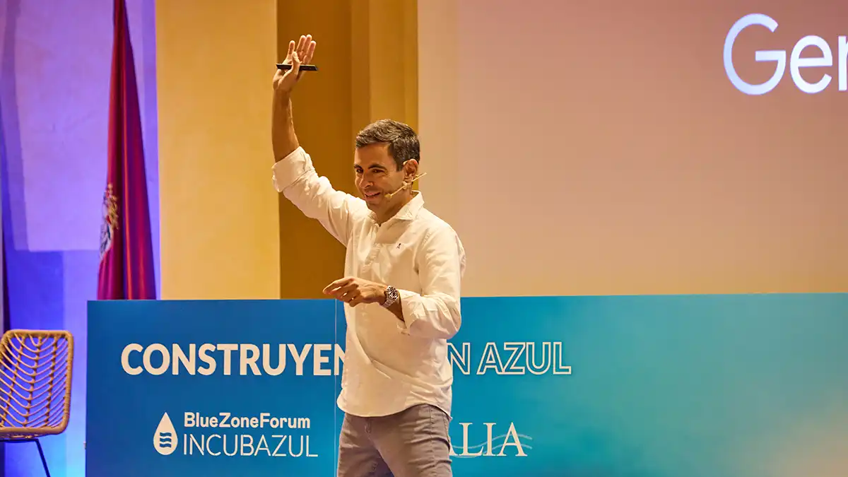 Javier Germade , un hombre de mediana edad, de pie, sonriente, levanta la mano en señal de saludo desde el escenario del Blue Zone Forum, con una pantalla de fondo proyectando la identidad visual del evento.