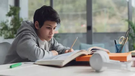 Chico estudiando delante de libros, toma notas en un cuaderno.