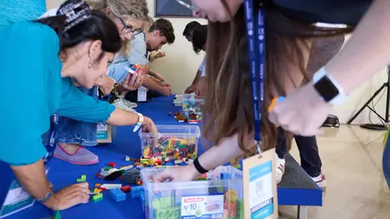 Varios asistentes sentados alrededor de mesas construyen figuras con piezas de LEGO de colores durante un taller práctico en el Blue Zone Forum 2025.