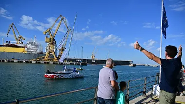 El primer velero de la Globe 40 entra en la Bahía de Cádiz mientras varias personas lo reciben desde el Paseo de Astilleros con saludos