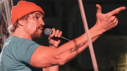 El cantante Macaco en el escenario, con el micrófono en una mano y la otra extendida hacia delante, señalando al público en un gesto característico.